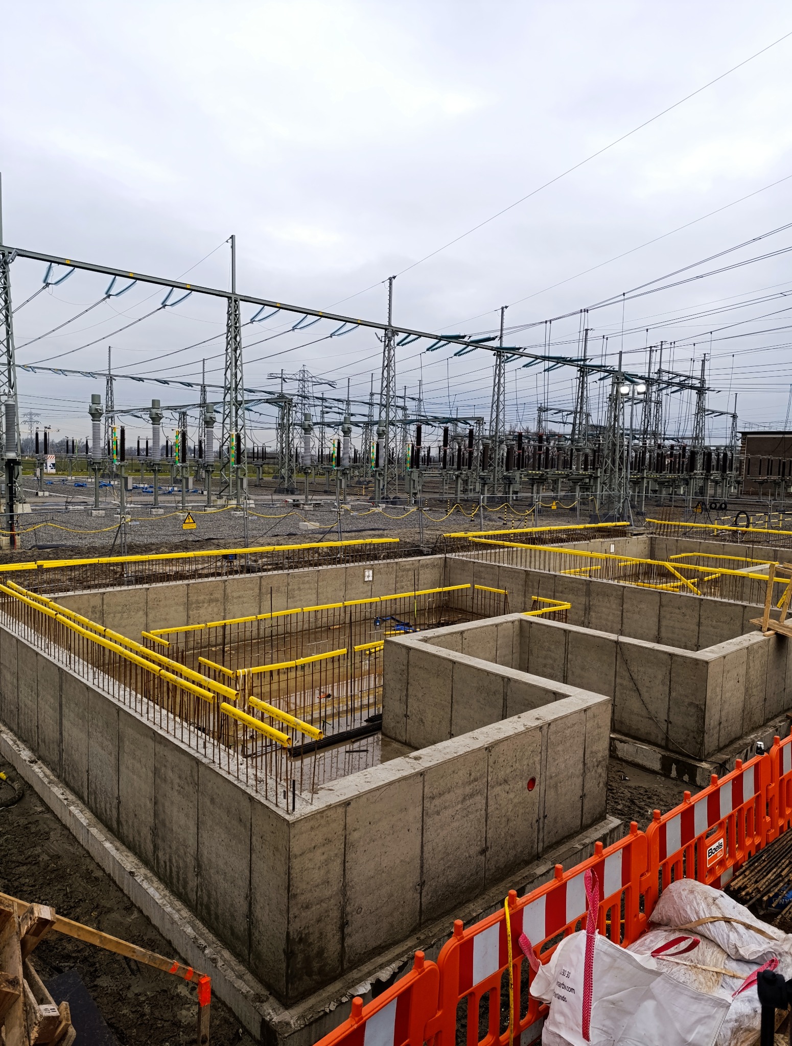 Betonowe fundamenty pod budowę stacji transformatorowej, z żółtymi barierkami ochronnymi i widoczną konstrukcją stalową w tle, wykonane w pochmurny dzień.