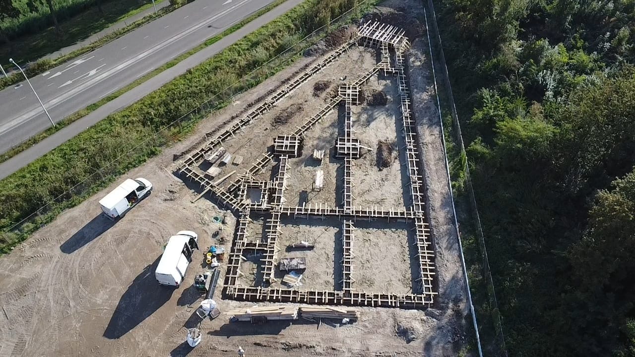 Widok z drona na drewniane szalunki fundamentów domu jednorodzinnego, z dwoma białymi samochodami dostawczymi obok placu budowy.