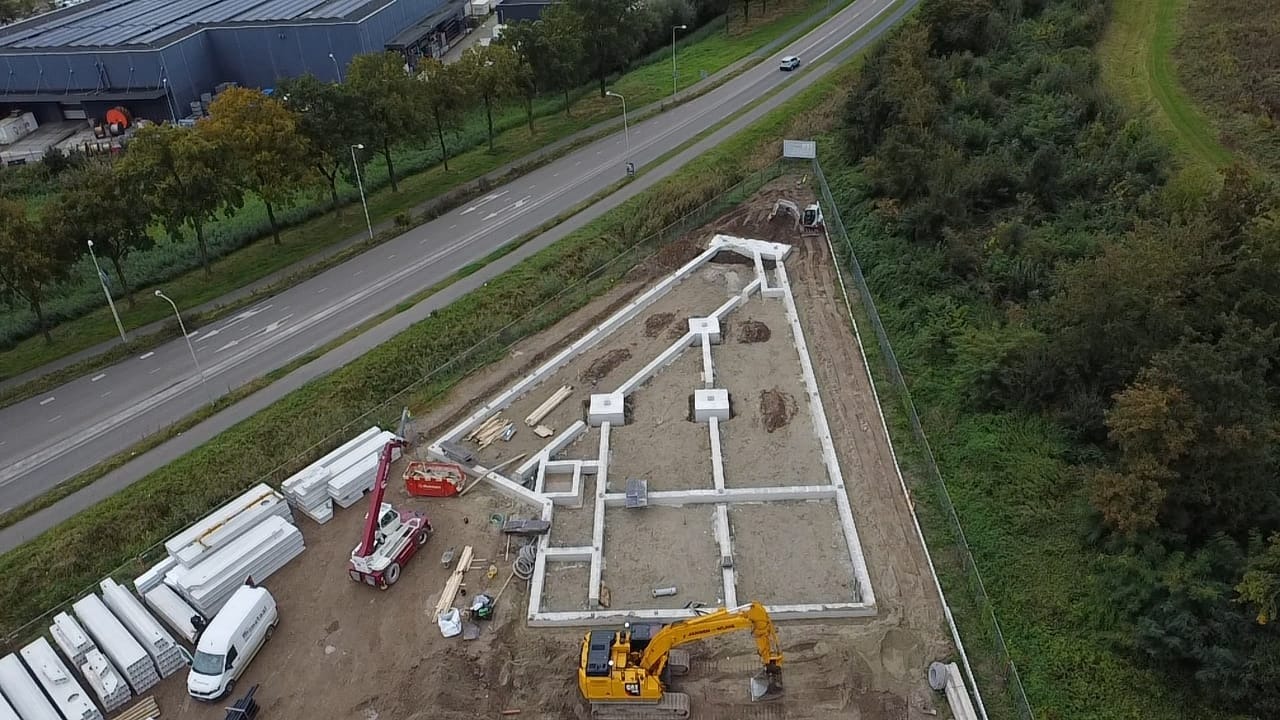 Fundamenty domu w kształcie trapezu, widok z góry. Na budowie stoi żółta koparka, a obok leżą elementy konstrukcyjne. W tle droga i drzewa.