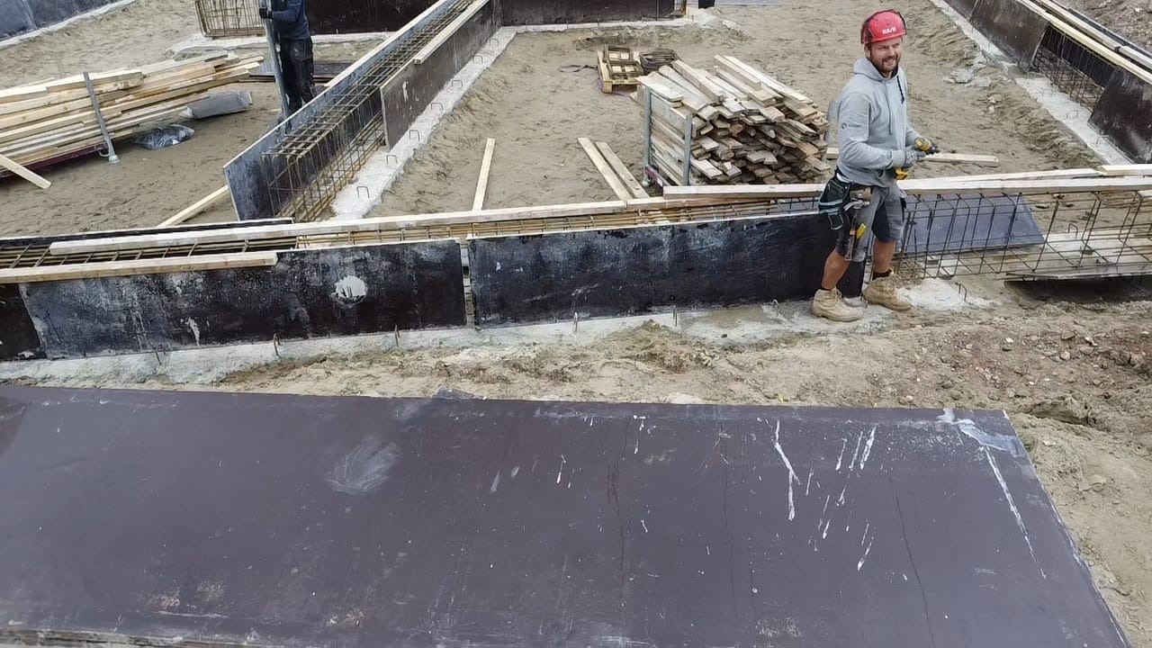 Budowa fundamentów: szalunek zbrojony stalą, pracownik w kasku. Widoczne deski i narzędzia. Etap przygotowawczy fundamentów pod budynek.