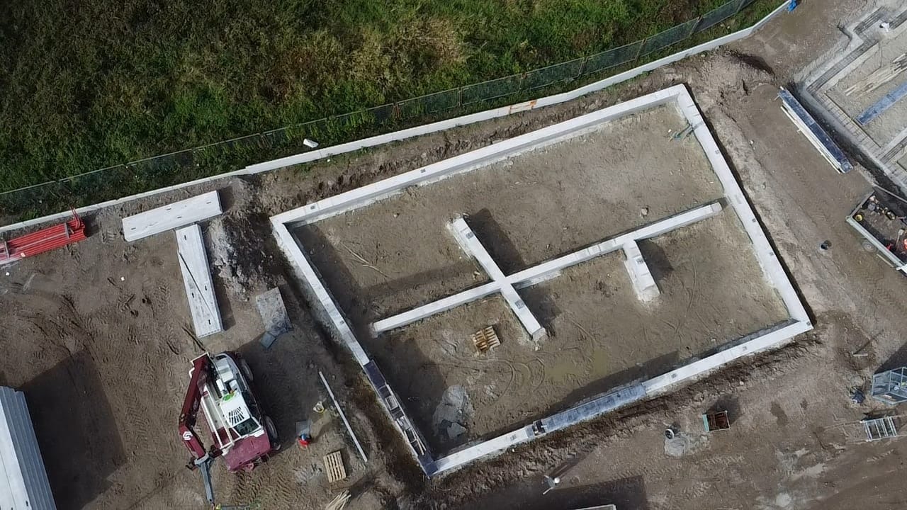 Widok z drona na wylane fundamenty pod budynek z zaznaczonym zbrojeniem krzyżowym, w tle trawa i częściowo widoczny sprzęt budowlany.