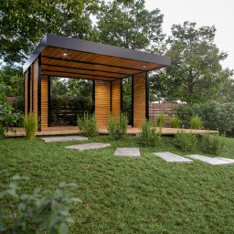 Modelarium - Wizualizacja nowoczesnej altany ogrodowej z drewnianą podłogą i ścieżką z kamiennych płyt na trawniku, otoczonej zielenią i drzewami. Oświetlenie wbudowane w sufit.