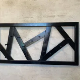 Czarna, metalowa konstrukcja dekoracyjna z nitami, zamontowana na szarej ścianie. Geometryczny wzór z ukośnych i pionowych elementów w prostokątnej ramie.