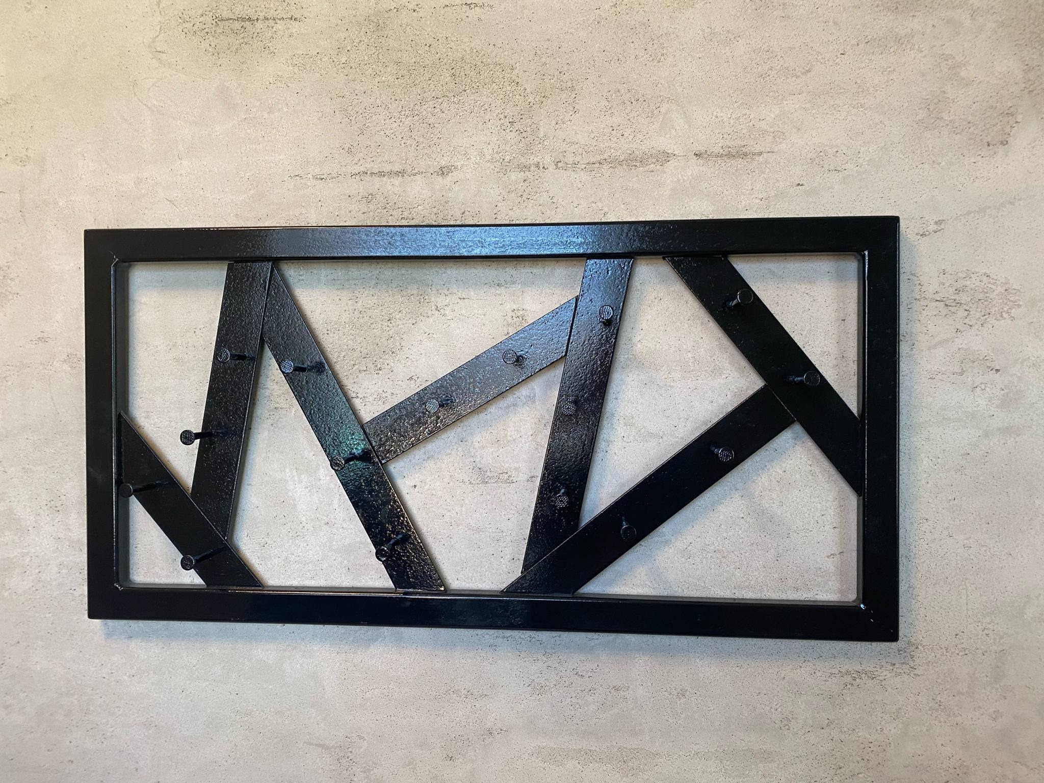 Czarna, metalowa konstrukcja dekoracyjna z nitami, zamontowana na szarej ścianie. Geometryczny wzór z ukośnych i pionowych elementów w prostokątnej ramie.