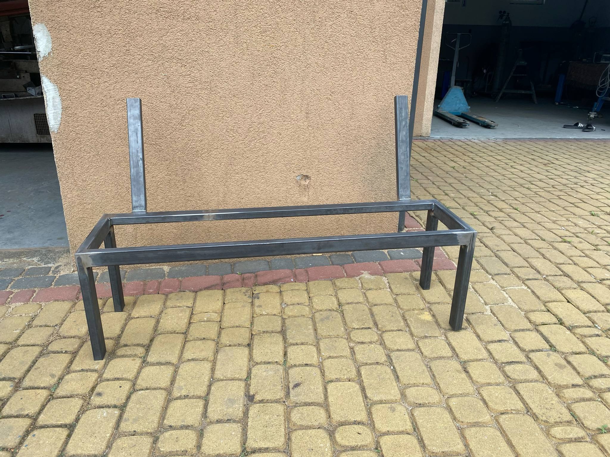 Metalowa konstrukcja z profili kwadratowych, prawdopodobnie rama ławki, ustawiona na kostce brukowej przed budynkiem z beżową elewacją, widok z boku.