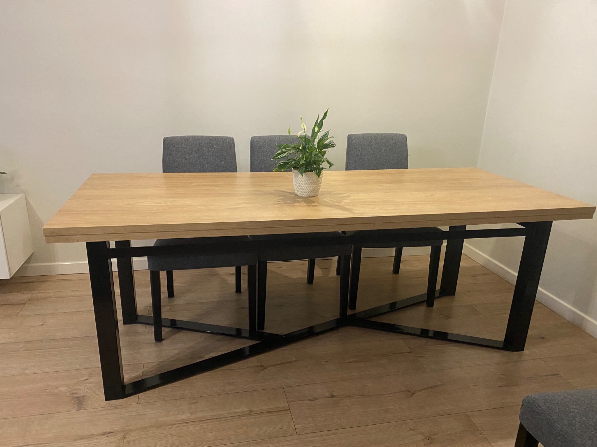 Stół z drewnianym blatem i czarną, metalową podstawą w kształcie krzyża. Na stole stoi doniczka z kwiatem. W tle trzy szare krzesła.