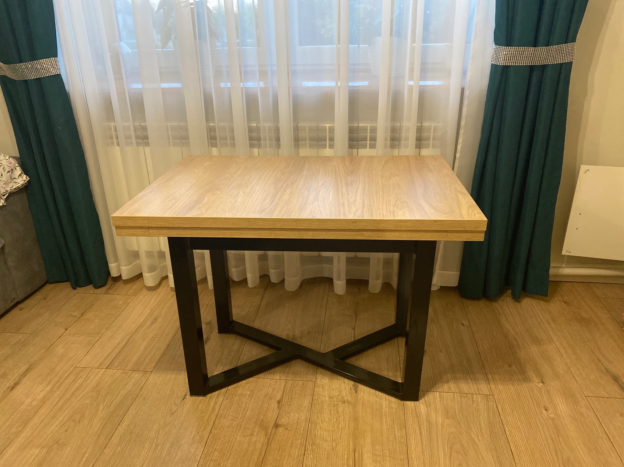 Stół z blatem imitującym jasne drewno i czarną, metalową podstawą w kształcie litery X, na tle okna z białymi firankami i zielonymi zasłonami.