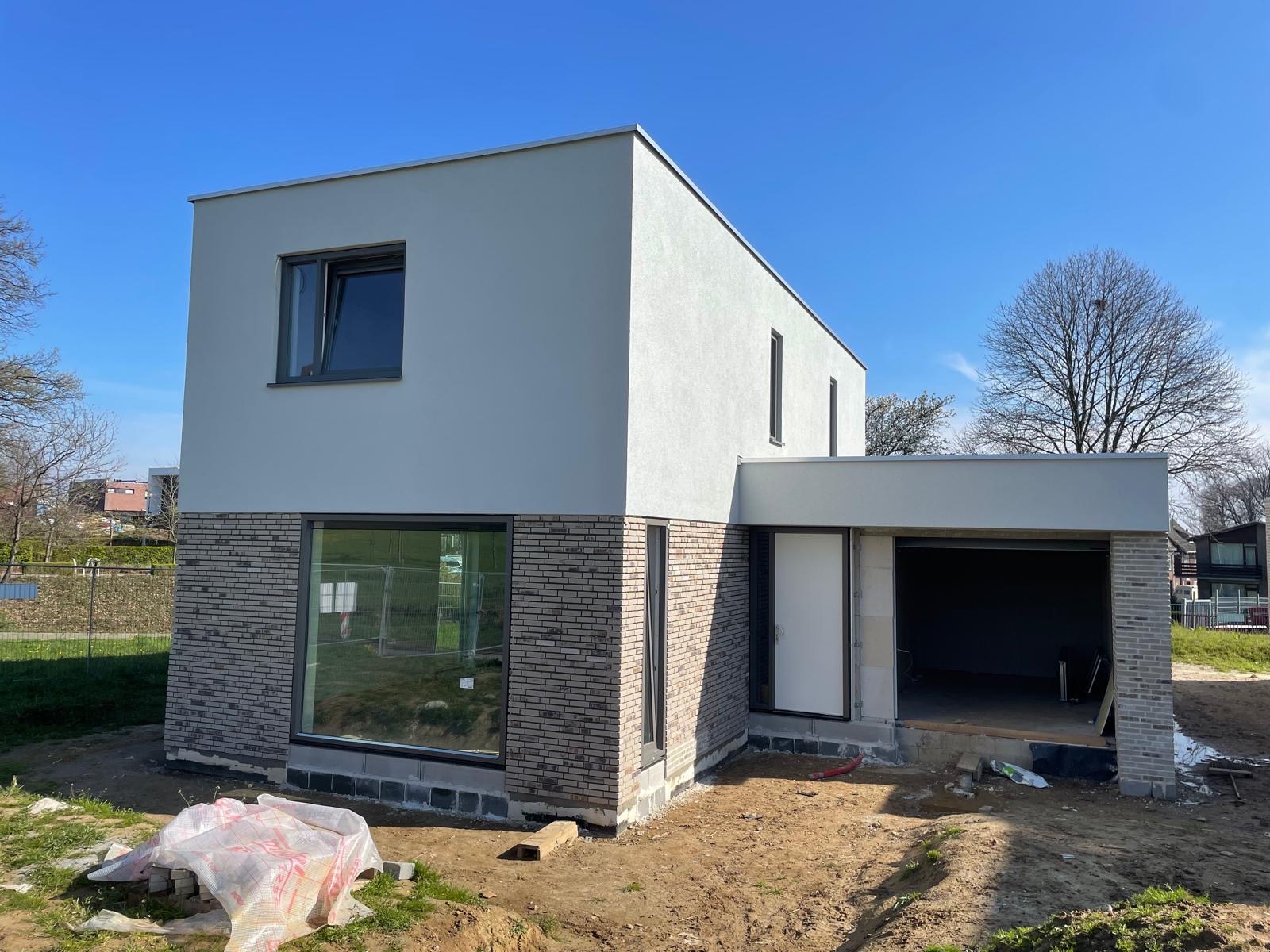 Nowoczesny dom z elewacją w dwóch odcieniach szarości, z wyraźnym podziałem na część tynkowaną i ceglaną. Widoczny fragment ogrodu i błękitne niebo.