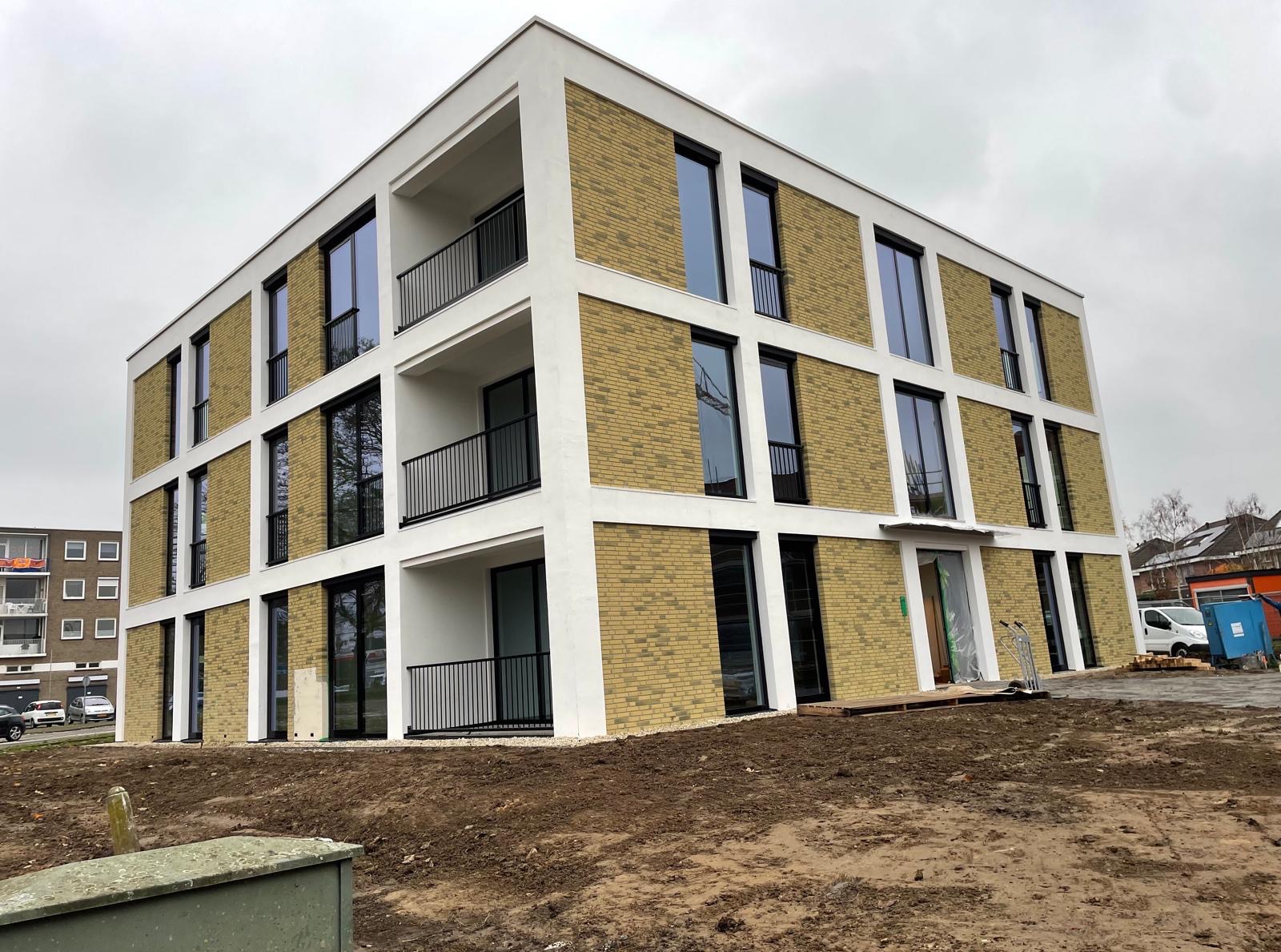 Nowoczesny budynek z elewacją z żółtej cegły i białymi ramami okiennymi. Widoczne balkony z czarnymi balustradami. Teren wokół budynku jest jeszcze w trakcie prac wykończeniowych.