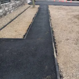 Nowa, czarna nawierzchnia asfaltowa z obrzeżami, otoczona żwirem i ziemią. W tle widoczny sprzęt budowlany i słup energetyczny. Prace drogowe w toku.