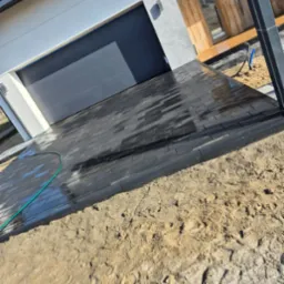 Ułożona, czarna kostka brukowa na podjeździe przed nowoczesnym domem z szarą bramą garażową i drewnianym wykończeniem, częściowo na piasku, z zielonym wężem ogrodowym.