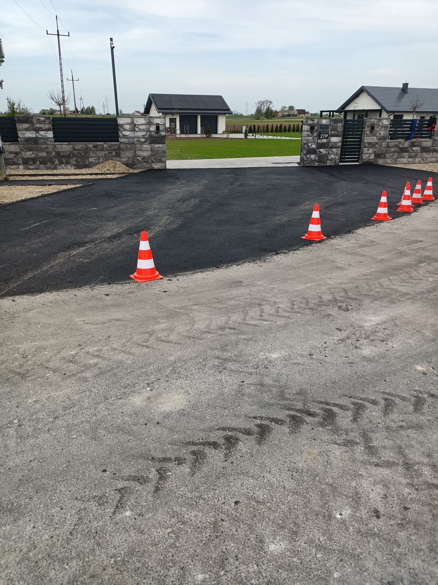 Świeżo wylany asfalt na podjeździe domu jednorodzinnego, wygrodzony pomarańczowymi pachołkami. Widok na nowoczesną zabudowę przez bramę z betonowych bloczków.