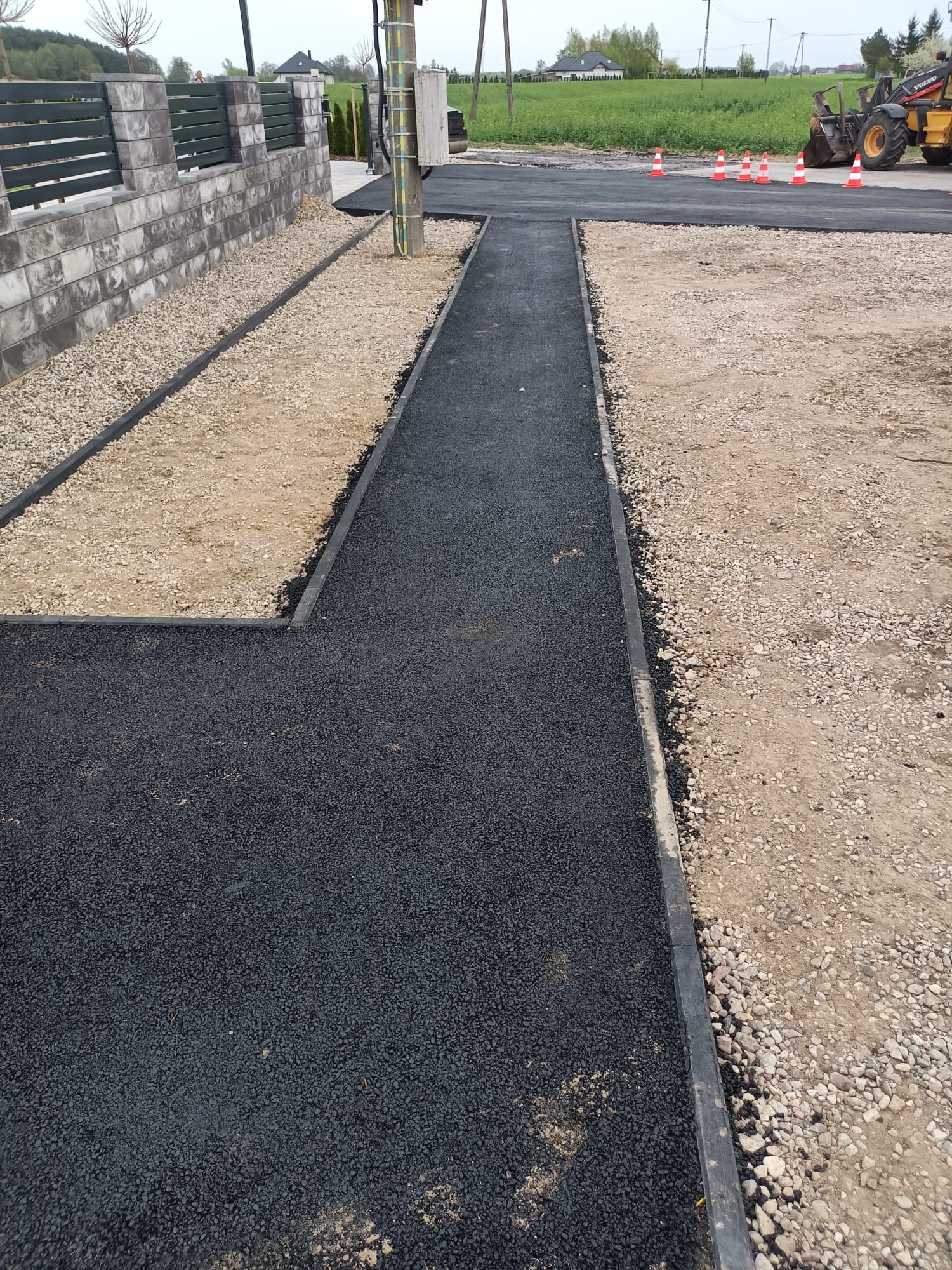 Nowa, czarna nawierzchnia asfaltowa z obrzeżami, otoczona żwirem i ziemią. W tle widoczny sprzęt budowlany i słup energetyczny. Prace drogowe w toku.