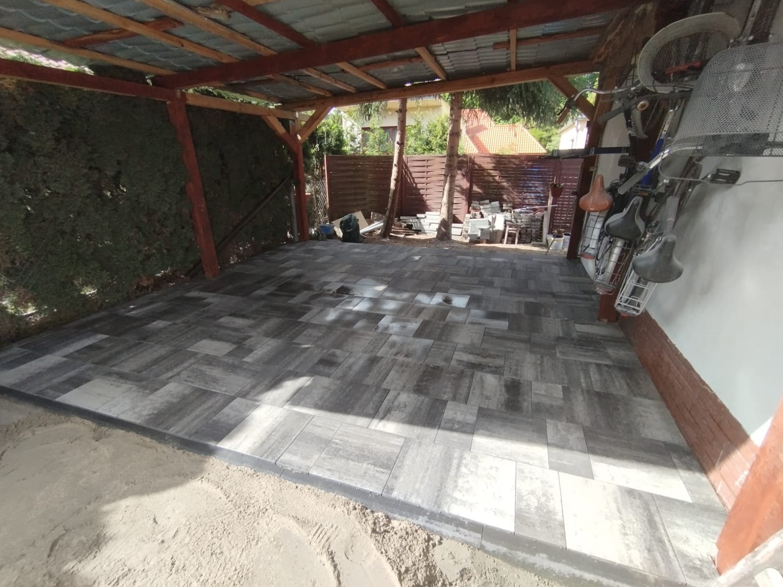 Zadaszona wiata z nowo ułożoną, szarą kostką brukową o nieregularnym wzorze. W tle widoczne przygotowane kostki oraz fragment roweru oparty o ścianę.