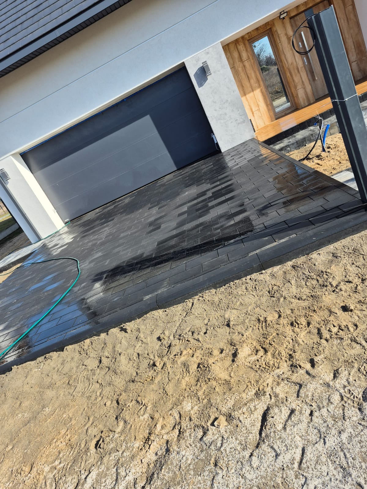 Ułożona, czarna kostka brukowa na podjeździe przed nowoczesnym domem z szarą bramą garażową i drewnianym wykończeniem, częściowo na piasku, z zielonym wężem ogrodowym.