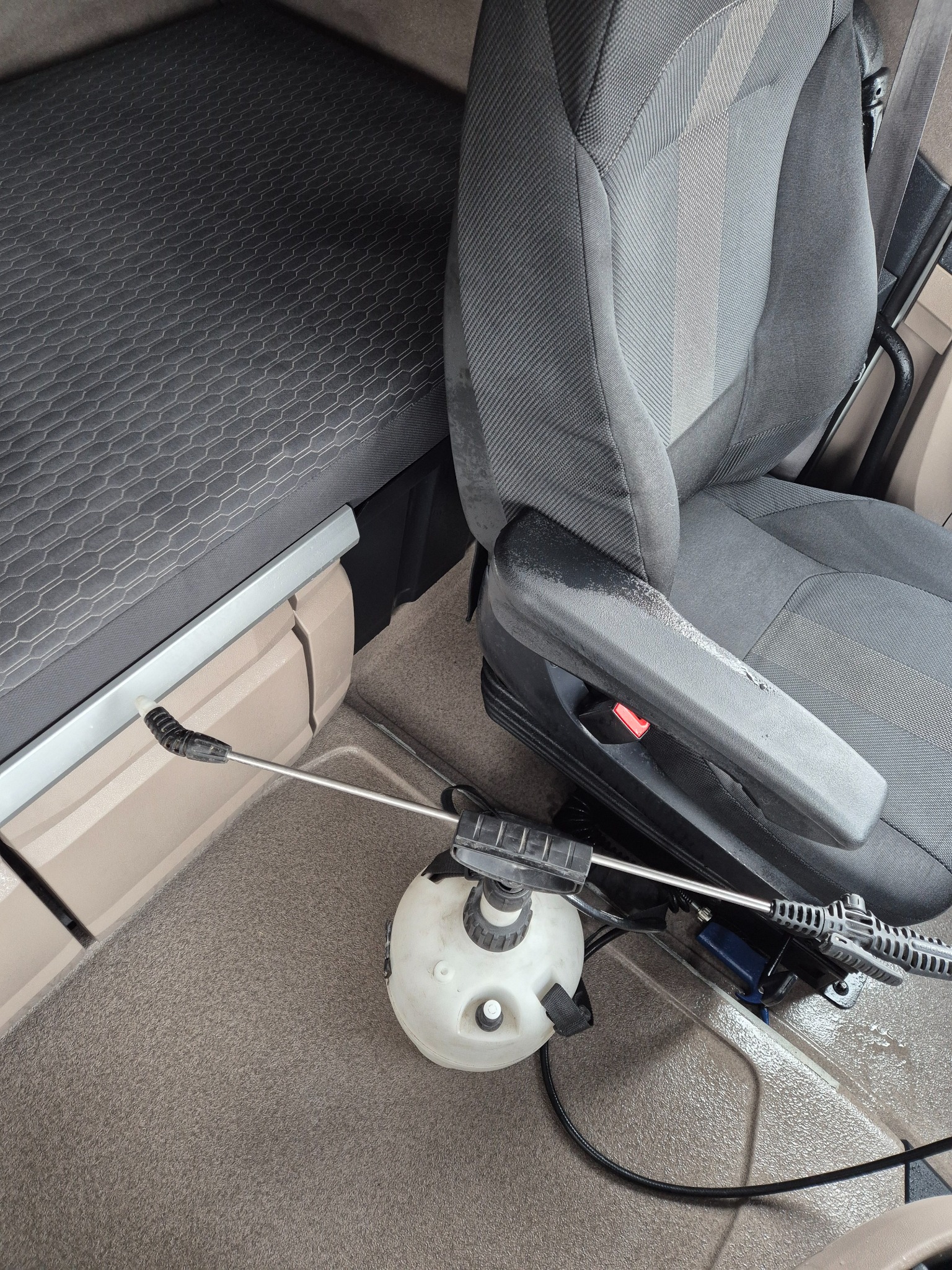 Wnętrze kabiny pojazdu z fotelem i leżanką, spryskiwacz ciśnieniowy na podłodze, prawdopodobnie w trakcie dezynfekcji lub czyszczenia.