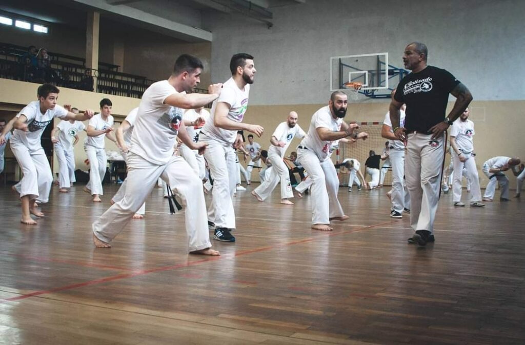 Trening capoeiry w Poznaniu: grupa osób w białych strojach ćwiczy pod okiem instruktora na sali gimnastycznej z parkietem i koszem do koszykówki w tle.