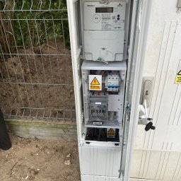 LUKON KONRAD UZARSKI - Otwarta skrzynka elektryczna z licznikiem i zabezpieczeniami, widoczne ostrzeżenia o napięciu. Na zewnątrz ogrodzenie z siatki i ziemia.