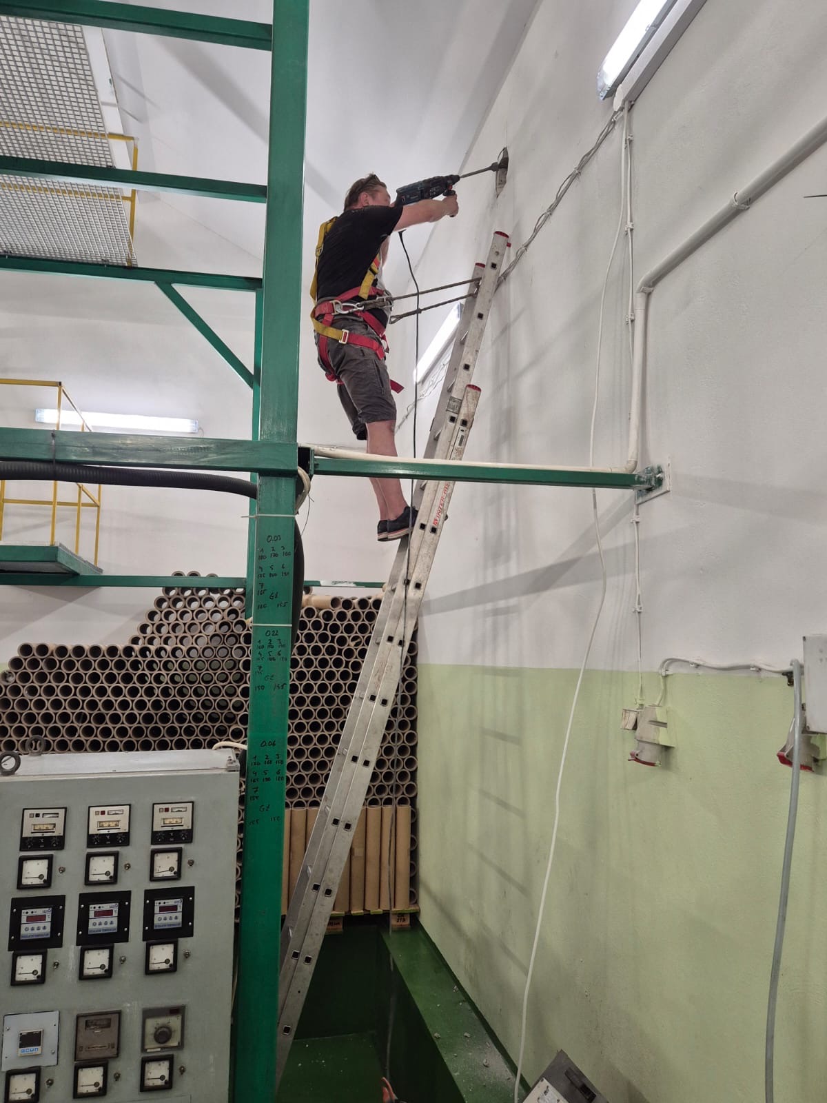 Elektryk w uprzęży na drabinie wierci otwór w ścianie. Widoczne kable, rury, skrzynka elektryczna. Praca na wysokości w hali przemysłowej.