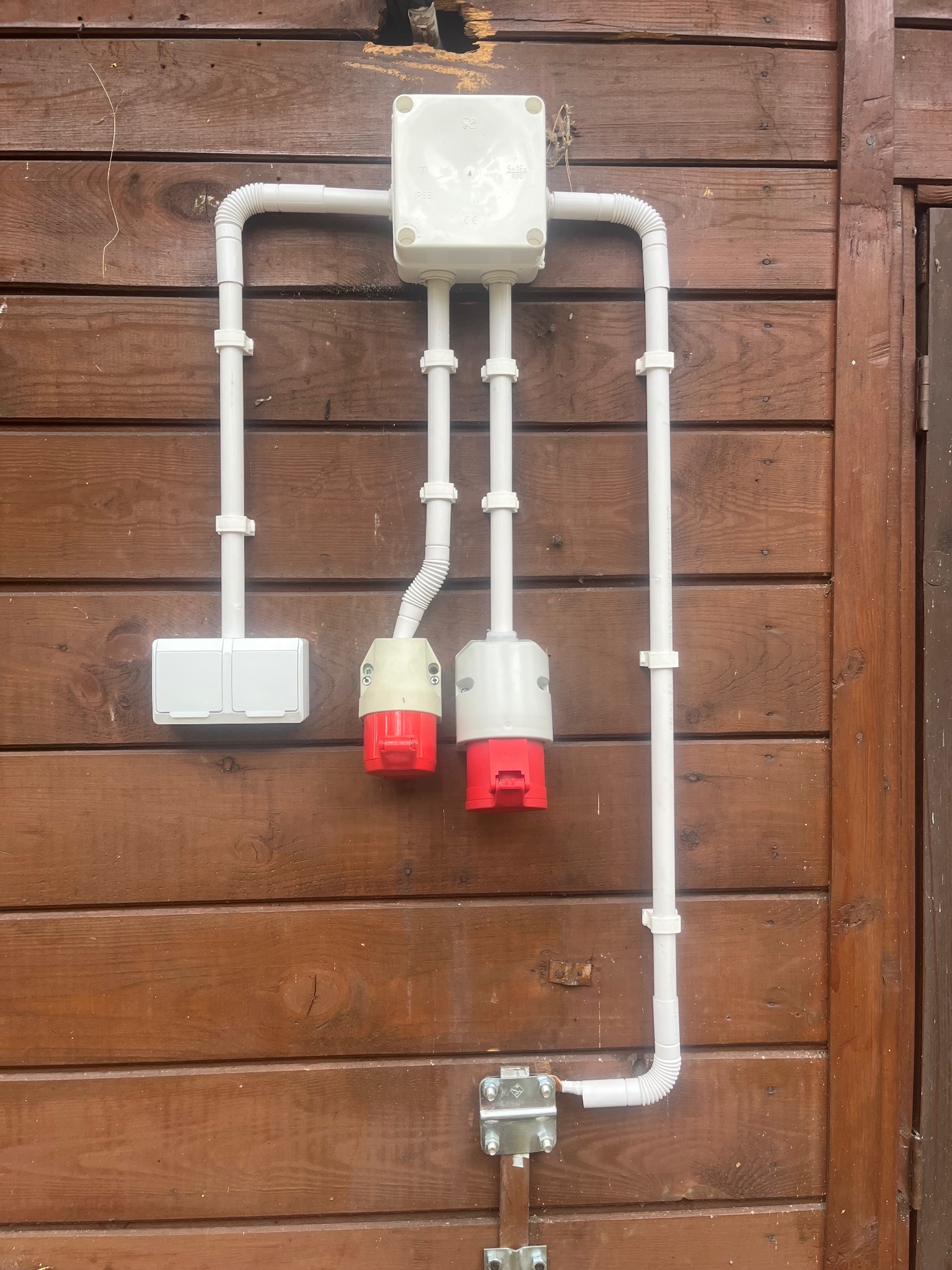 Białe przewody i gniazda elektryczne na brązowej, drewnianej ścianie. Widoczne kable w peszlu, puszka rozgałęźna i gniazda siłowe w kolorze białym z czerwonymi elementami.