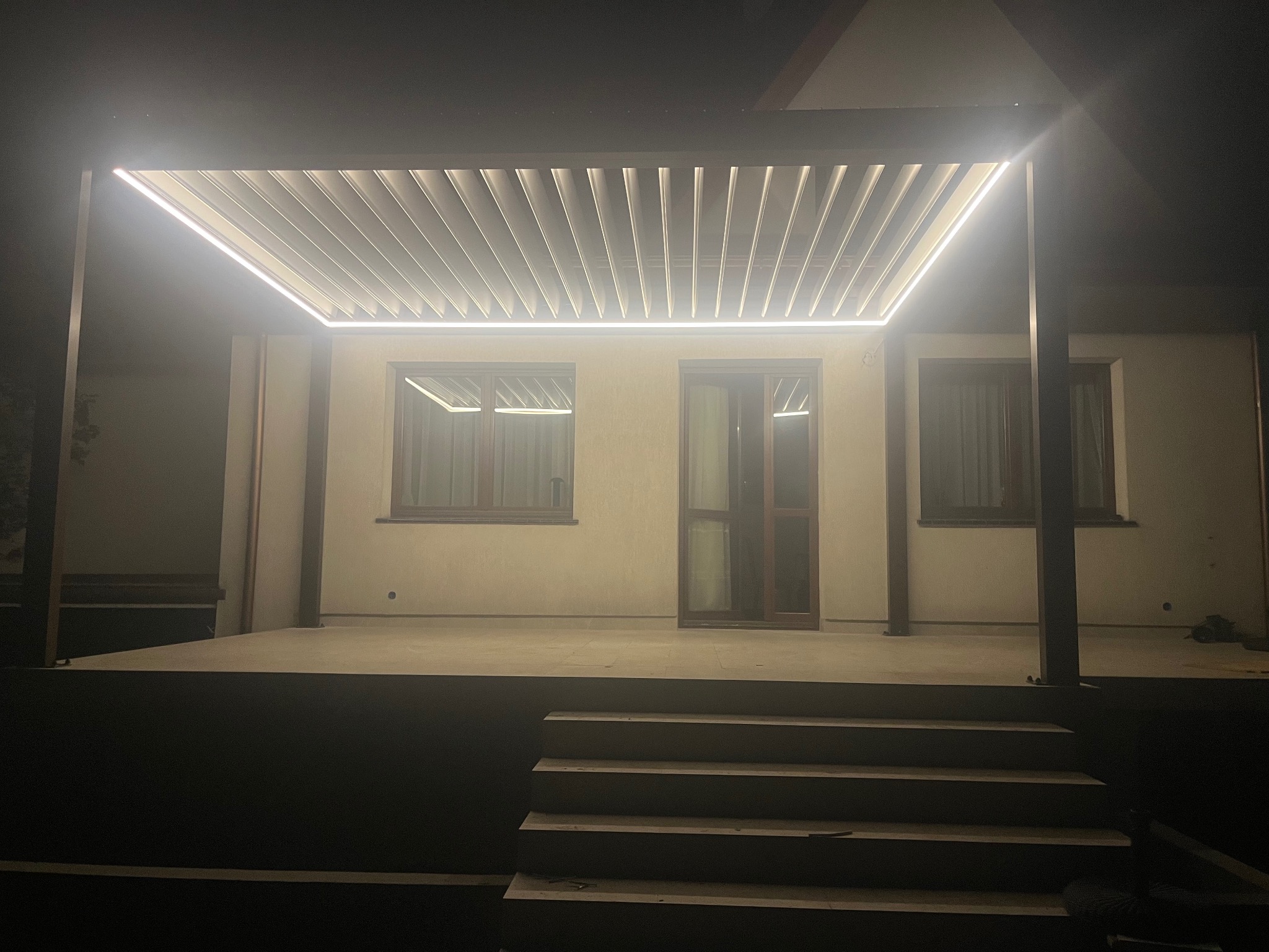 Oświetlona pergola z nowoczesnym, listwowym zadaszeniem i ledowym oświetleniem, widoczna nocą. Schody prowadzą na podest.
