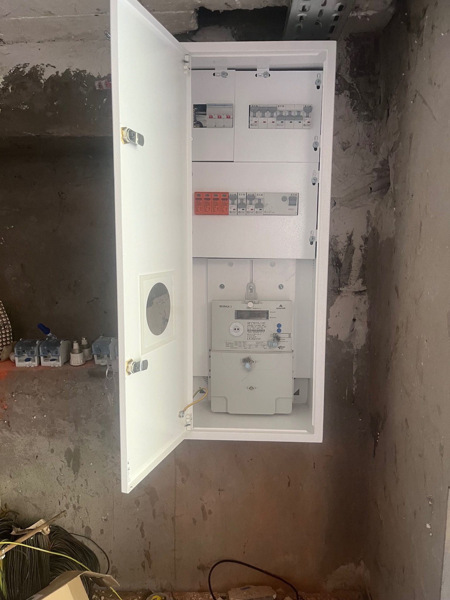 Otwarta skrzynka z nowym licznikiem i bezpiecznikami, zamontowana w surowym wnętrzu w Józefowie. Widoczne przewody i elementy instalacji.