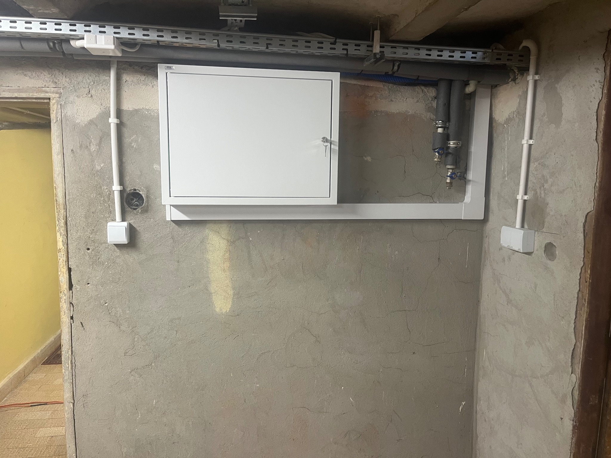 Biała skrzynka elektryczna z kluczem na surowej ścianie, szara rura instalacyjna pod sufitem. Widoczna instalacja elektryczna i wodna.