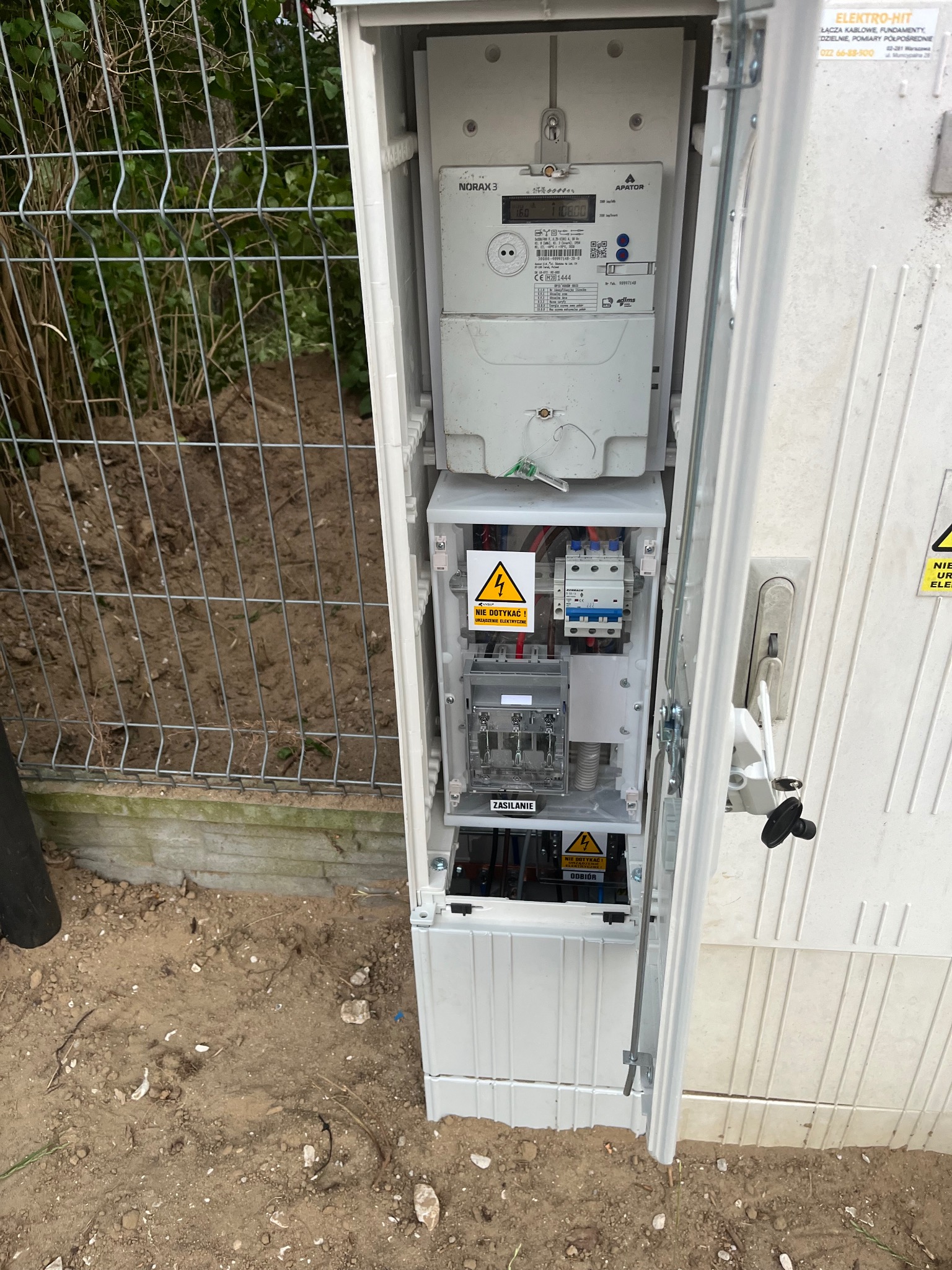Otwarta skrzynka elektryczna z licznikiem i zabezpieczeniami, widoczne ostrzeżenia o napięciu. Na zewnątrz ogrodzenie z siatki i ziemia.