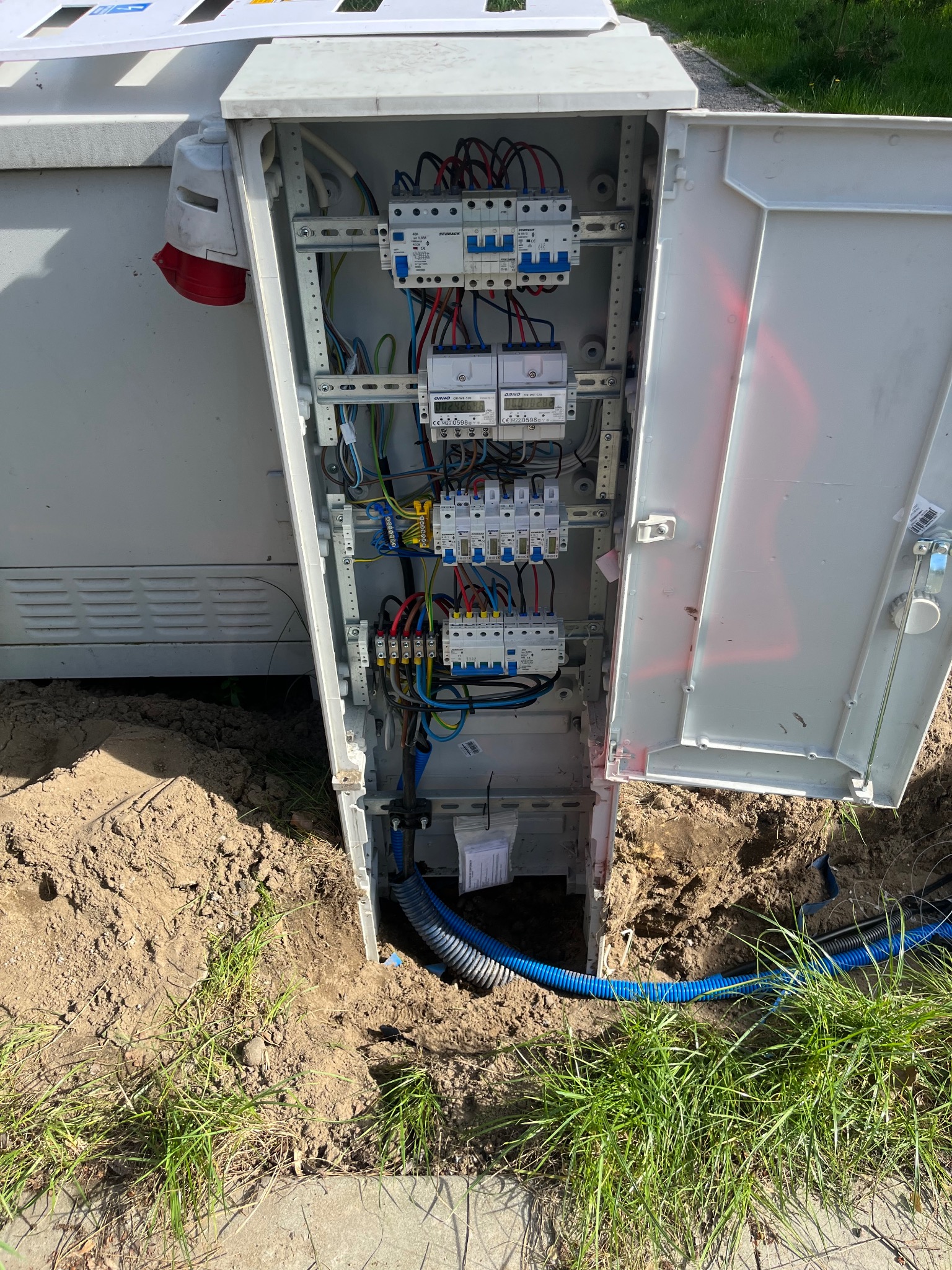 Otwarta skrzynka elektryczna z bezpiecznikami i przewodami, umieszczona w ziemi obok trawy. Widoczne niebieskie rury kablowe.