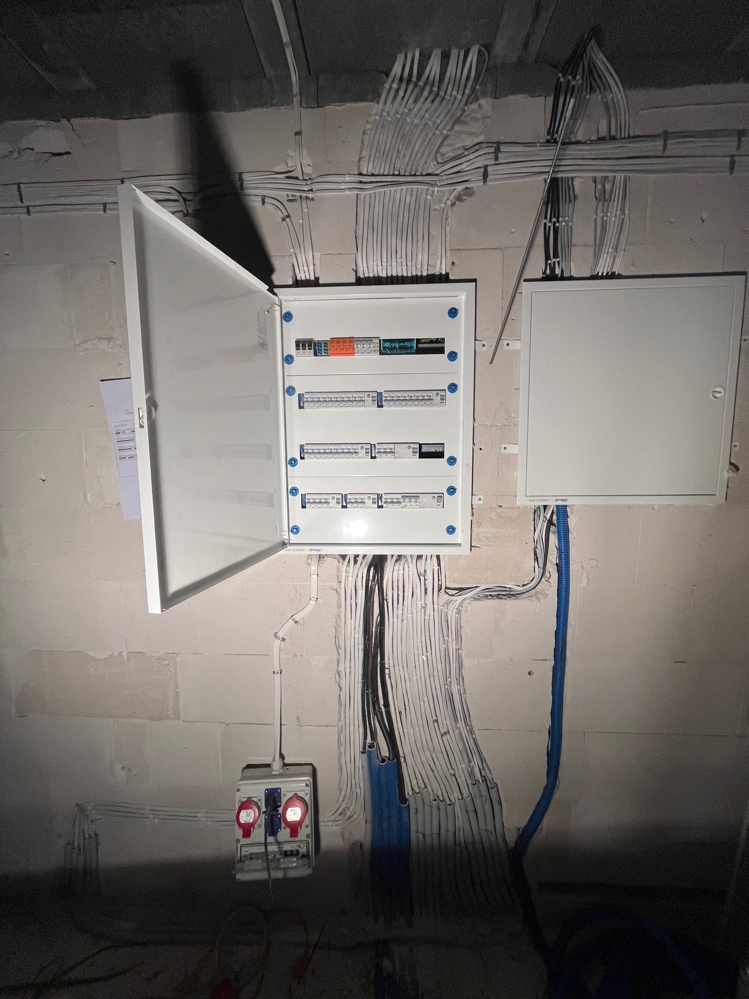 Otwarta skrzynka elektryczna z widocznymi bezpiecznikami na surowej ścianie z licznymi kablami. Dodatkowe gniazdo siłowe i peszel. Instalacja w trakcie realizacji.