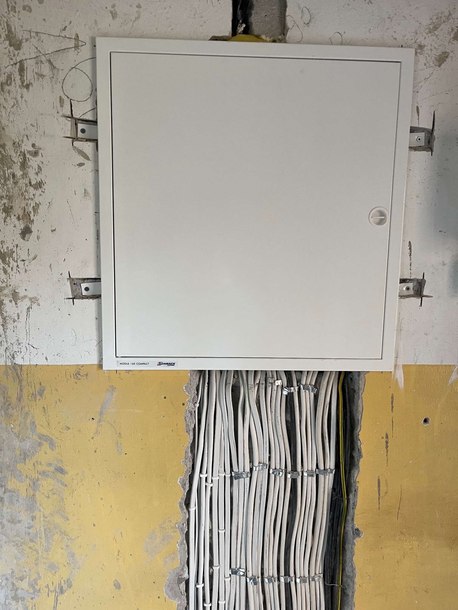 Biała skrzynka elektryczna Schrack zamontowana na surowej ścianie, z widocznymi kablami w korycie. Częściowo pomalowana ściana na żółto. Instalacja w trakcie prac.