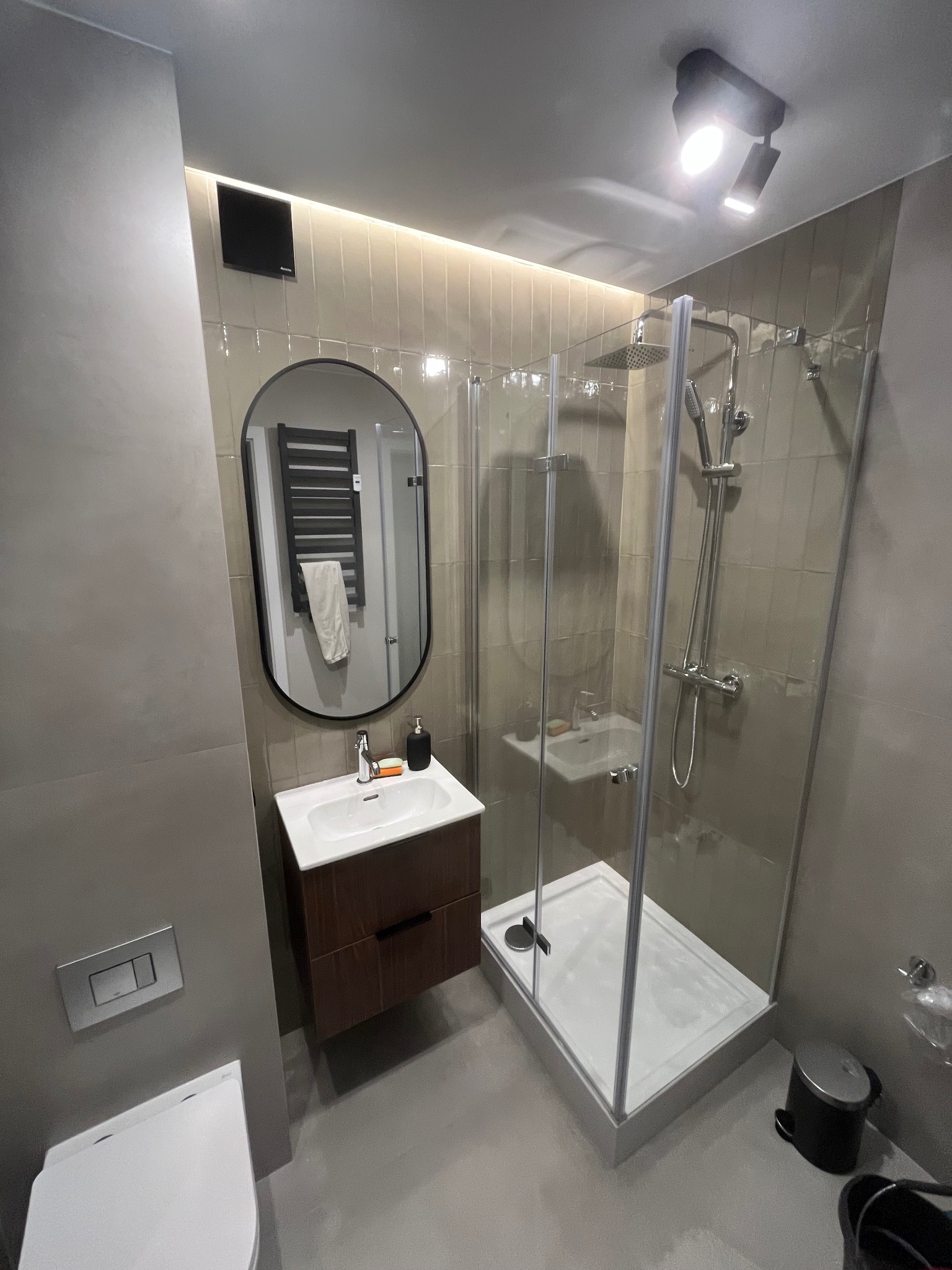 Nowoczesna łazienka z kabiną prysznicową, umywalką z szafką w kolorze drewna i owalnym lustrem. Jasne płytki, punktowe oświetlenie.