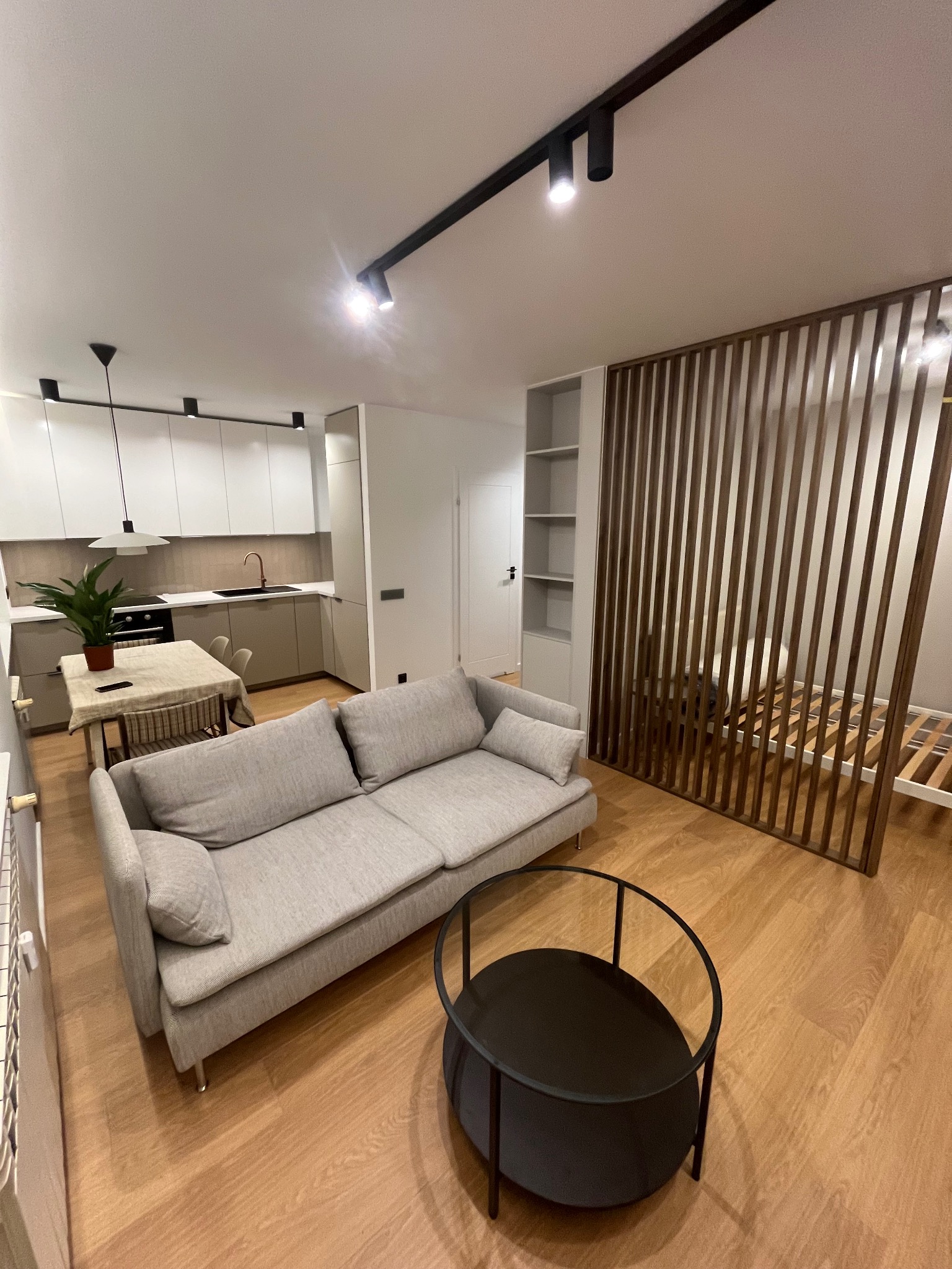 Nowoczesne oświetlenie szynowe z reflektorami w jasnym wnętrzu mieszkania z aneksem kuchennym, drewnianą ścianką działową i szarą kanapą.