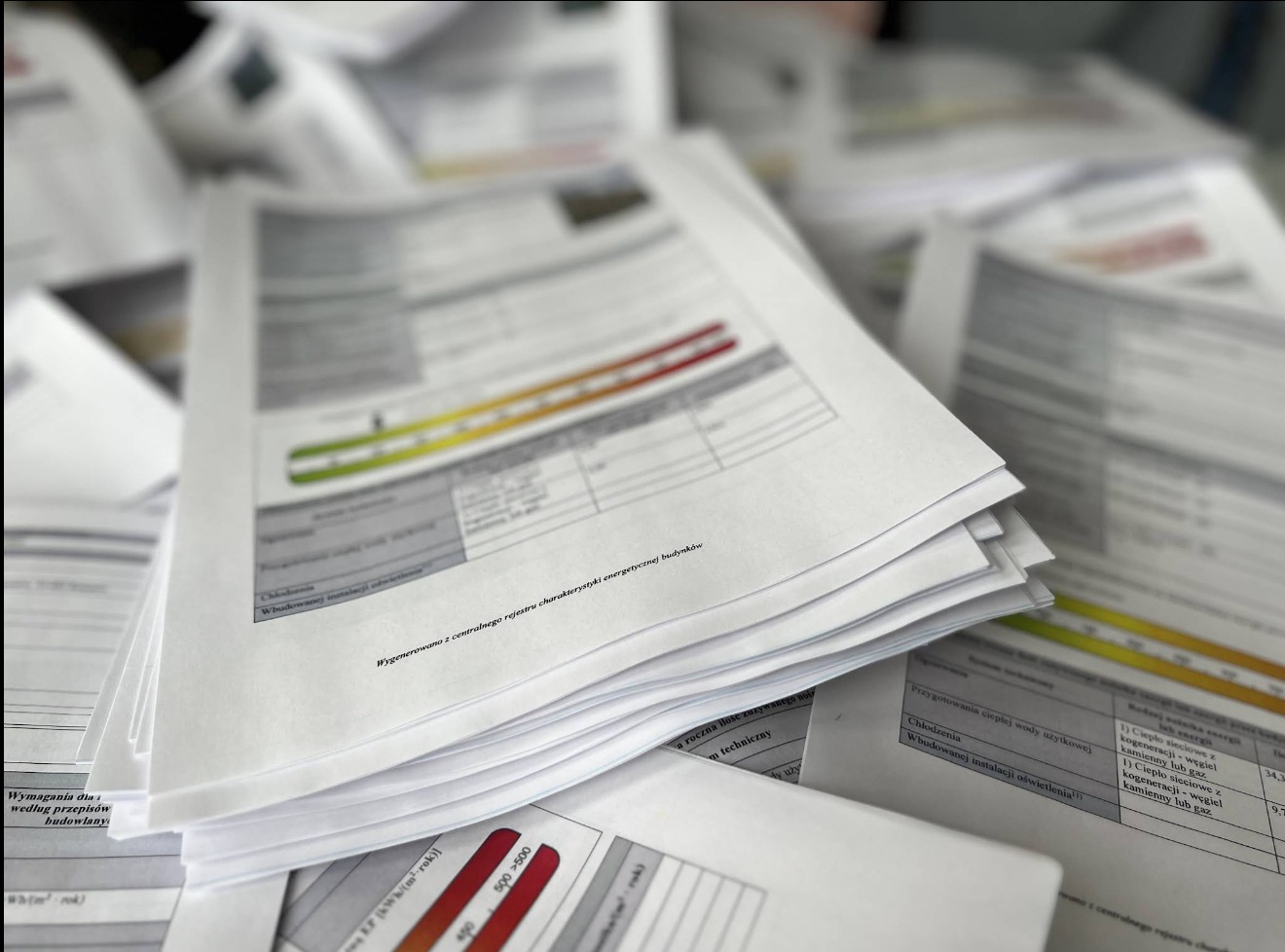 Stos dokumentów świadectw energetycznych ułożonych warstwami, z widocznymi wykresami i tabelami, w neutralnym oświetleniu biurowym.