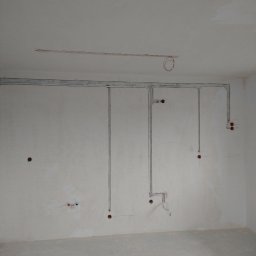 INSTALL ENERGY Łukasz Stępniak - Szare rurki instalacji elektrycznej na ścianie w surowym wnętrzu. Widoczne otwory pod gniazdka i puszki. Instalacja częściowo podtynkowa.