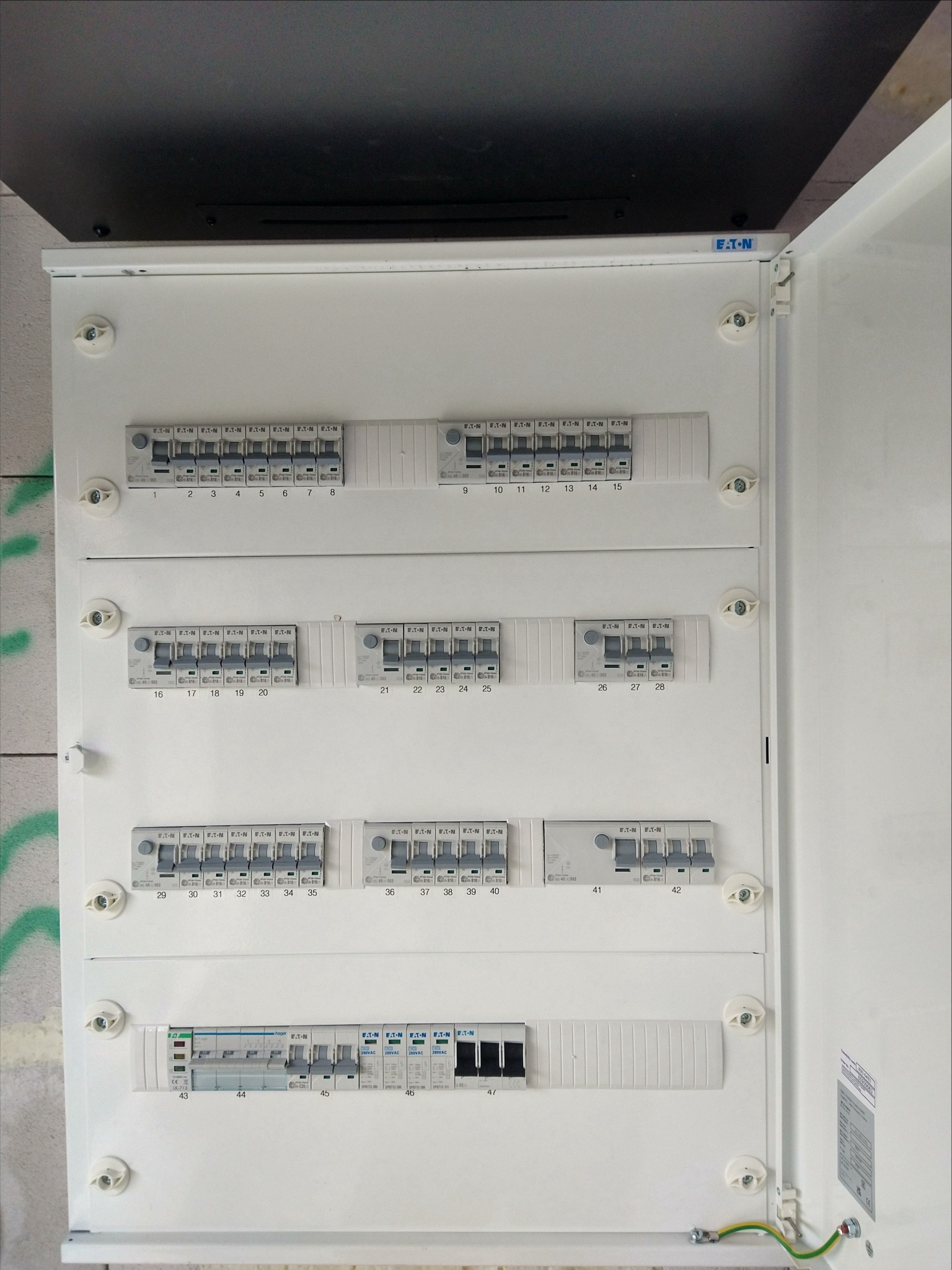 Otwarta biała skrzynka elektryczna z licznymi bezpiecznikami EATON i Hager, widoczne numery obwodów, przewód uziemiający.