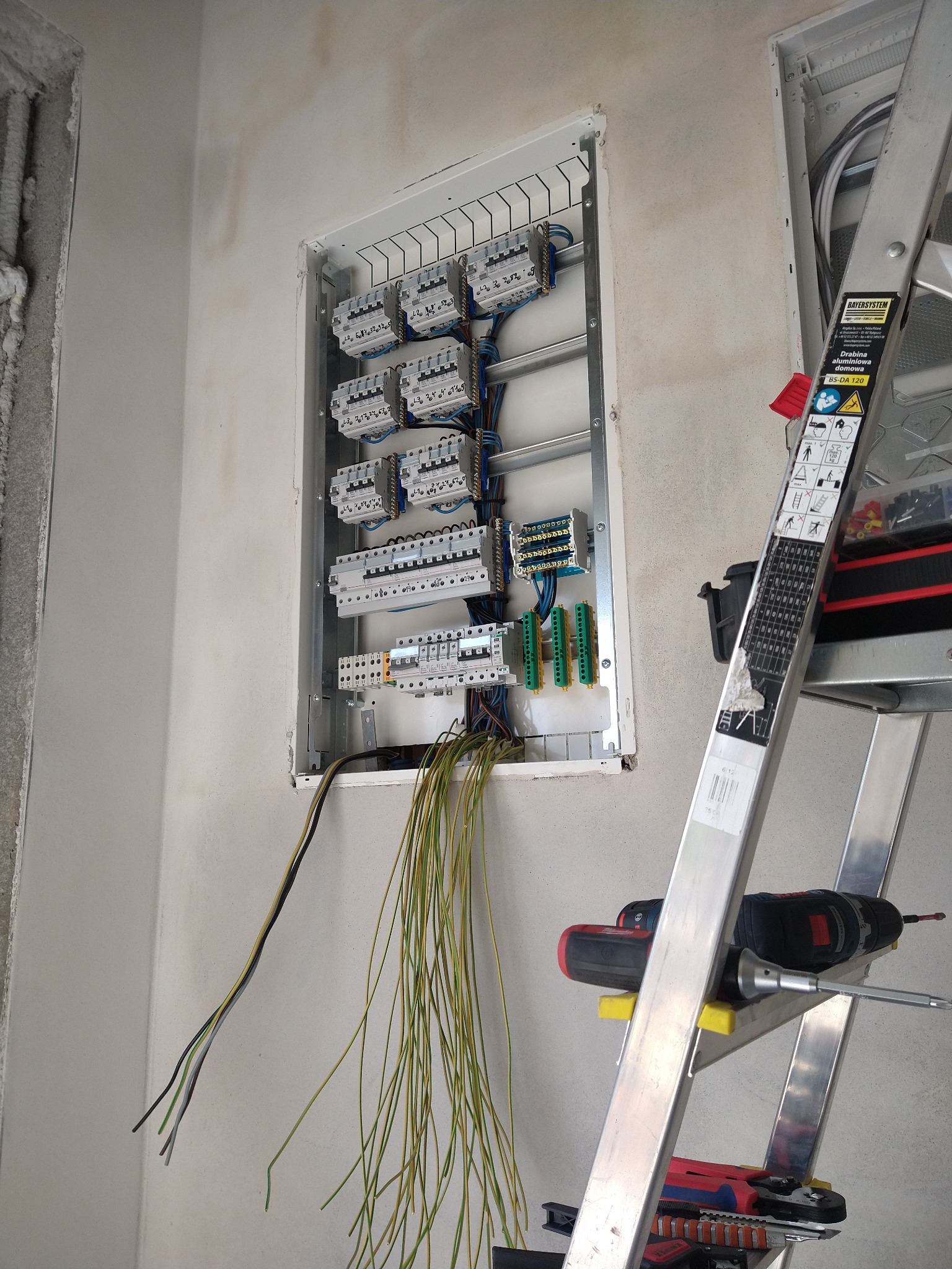 Rozdzielnica elektryczna w ścianie, widok z boku, z przewodami i drabiną. Widoczne narzędzia: wkrętarka i kombinerki. Ściana w stanie surowym.