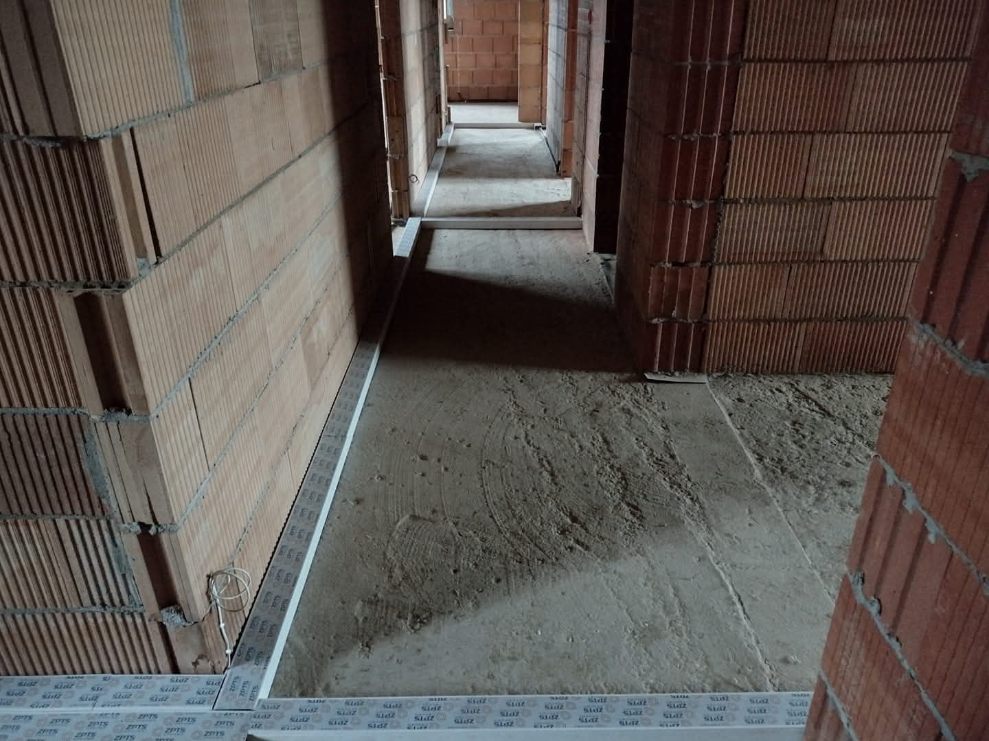 Wnętrze w trakcie budowy: surowy beton na podłodze, ściany z czerwonej cegły, widoczne elementy instalacji elektrycznej. Perspektywa długa, ukazująca kolejne pomieszczenia.