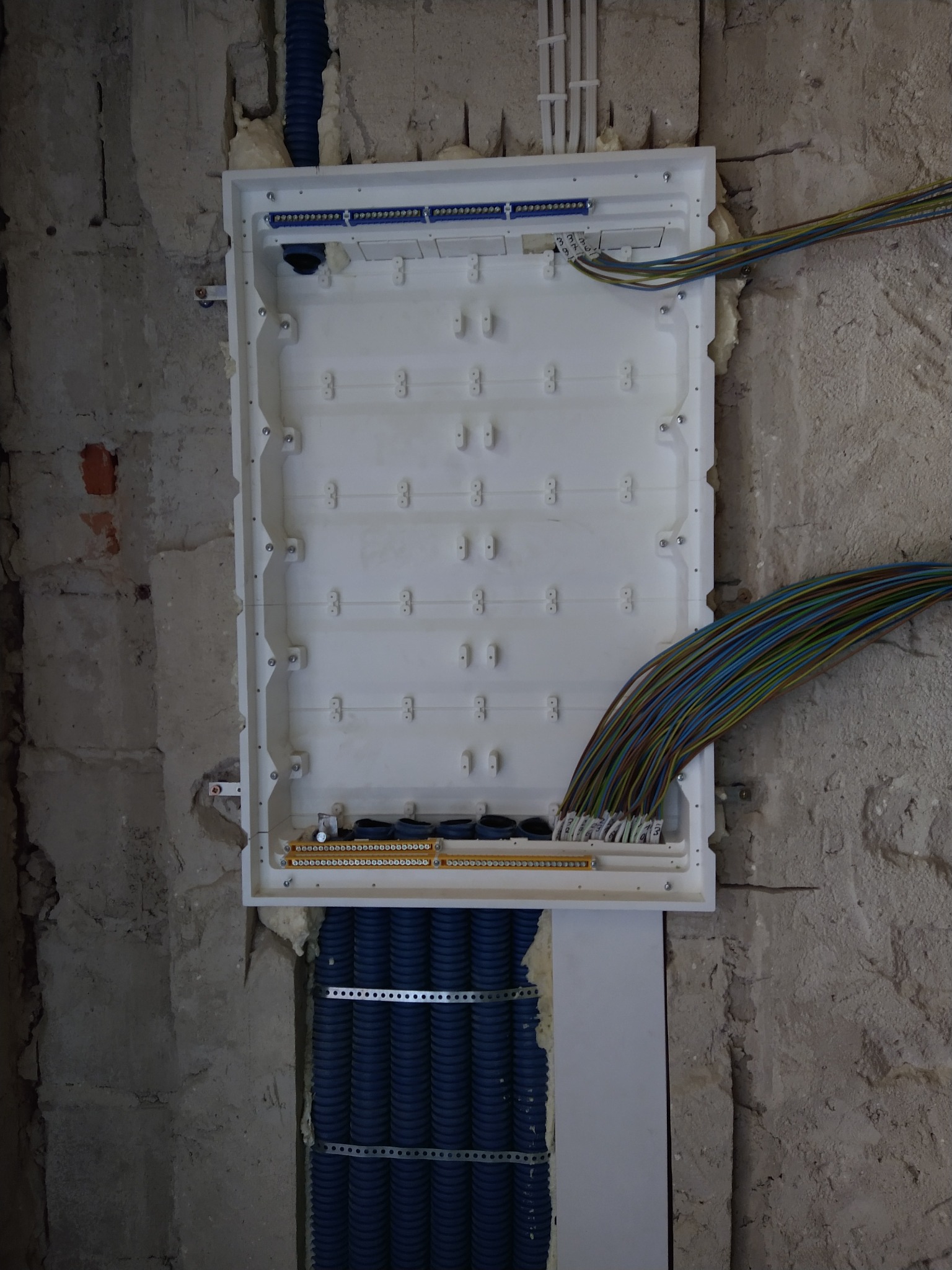 Rozdzielnia elektryczna natynkowa w trakcie montażu, widoczne kable w peszlach i przewody z oznaczeniami. Surowa ściana w tle. Etap prac instalacyjnych.