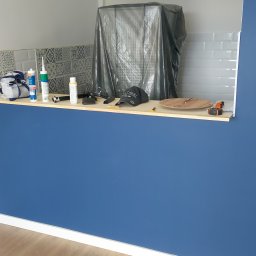 Damian Muchowski - Niebieska ściana po malowaniu z widocznymi narzędziami i materiałami na blacie; fragment kuchni z płytkami dekoracyjnymi.