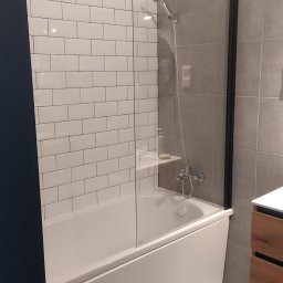 Damian Muchowski - Nowoczesna łazienka z wanną, przeszklonym parawanem z czarną ramą i białą armaturą. Ściany wyłożone białymi i szarymi płytkami. Minimalistyczny design.