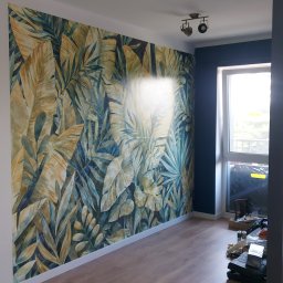 Damian Muchowski - Pokój po remoncie z fototapetą w motywy roślinne, drewnianą podłogą i ciemną ścianą przy oknie. Widoczne narzędzia malarskie.