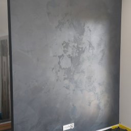 Damian Muchowski - Ściana z efektem dekoracyjnym w odcieniach szarości, zabezpieczona folią malarską i taśmą. Widoczne gniazdko elektryczne. Charakterystyczna faktura ściany.