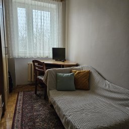 Damian Muchowski - Jasne wnętrze pokoju z beżowymi ścianami, drewnianym biurkiem i tapczanem z dekoracyjnymi poduszkami. Widok na okno z firanką i dywan w orientalne wzory.