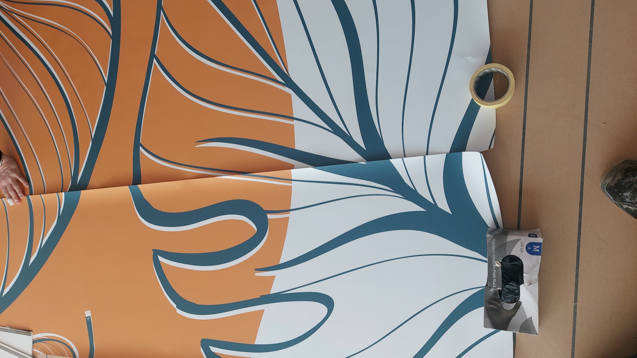 Nakładanie wzorzystej tapety w kolorach pomarańczowym i białym z niebieskimi akcentami. Widoczne narzędzia: taśma klejąca i rękawice ochronne. Praca w toku.