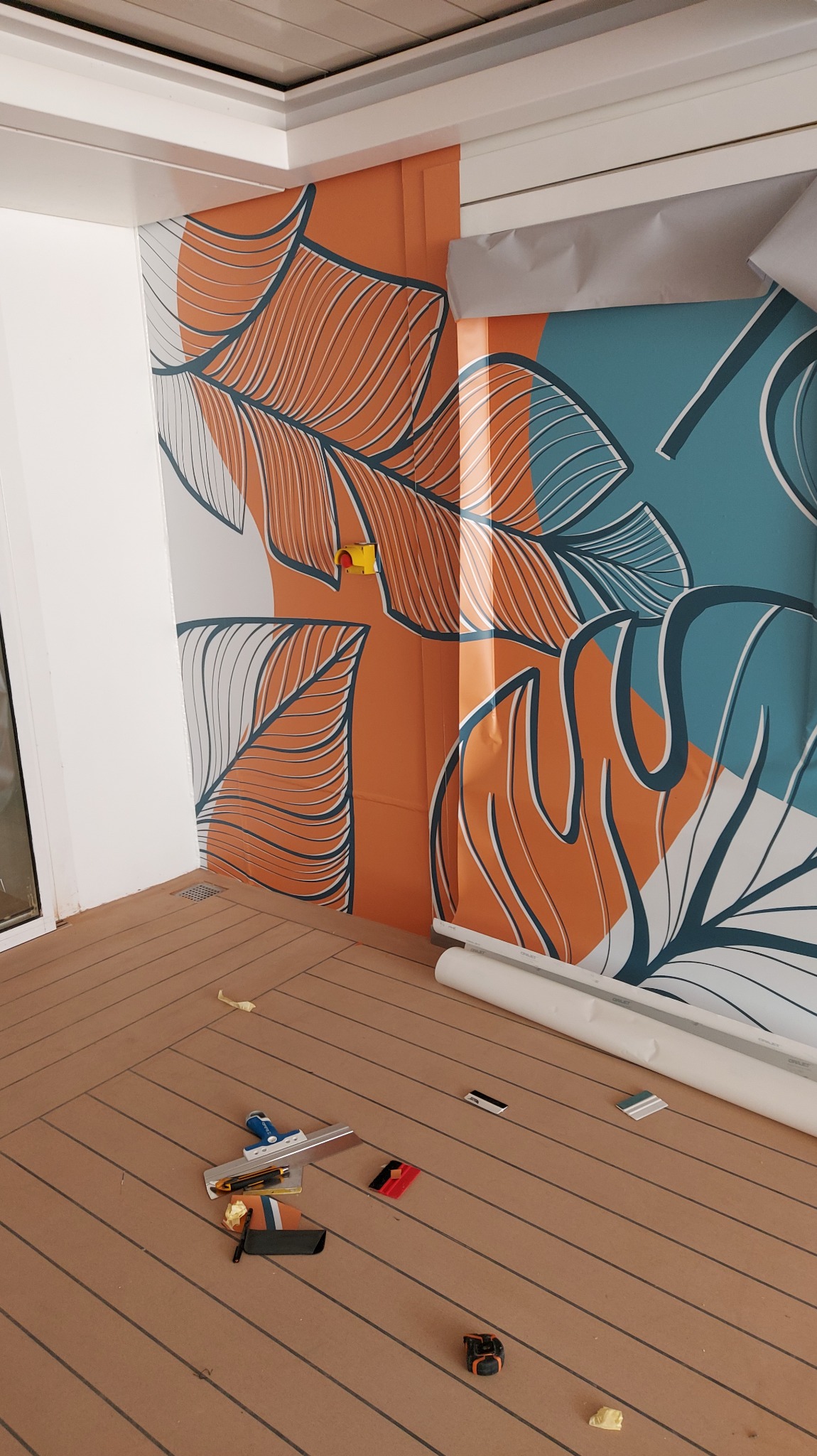 Naklejanie wzorzystej tapety z motywem liści na ścianę balkonu. Na podłodze narzędzia i resztki tapety. Widoczny fragment okna i sufitu.