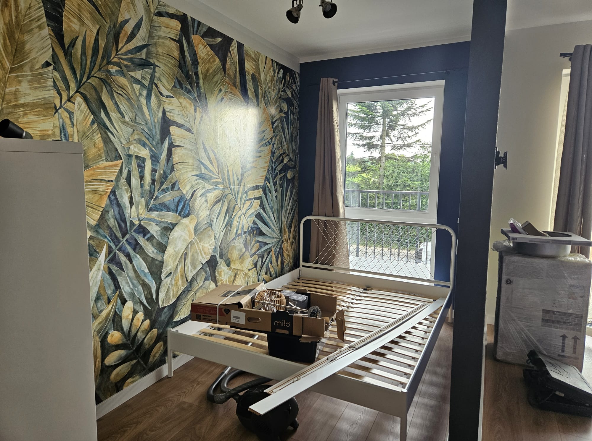 Pokój w trakcie urządzania: białe łóżko z materacem, kartony, odkurzacz, ściana z tapetą w motywy roślinne, okno z widokiem na zieleń. Etap wykończenia wnętrza.