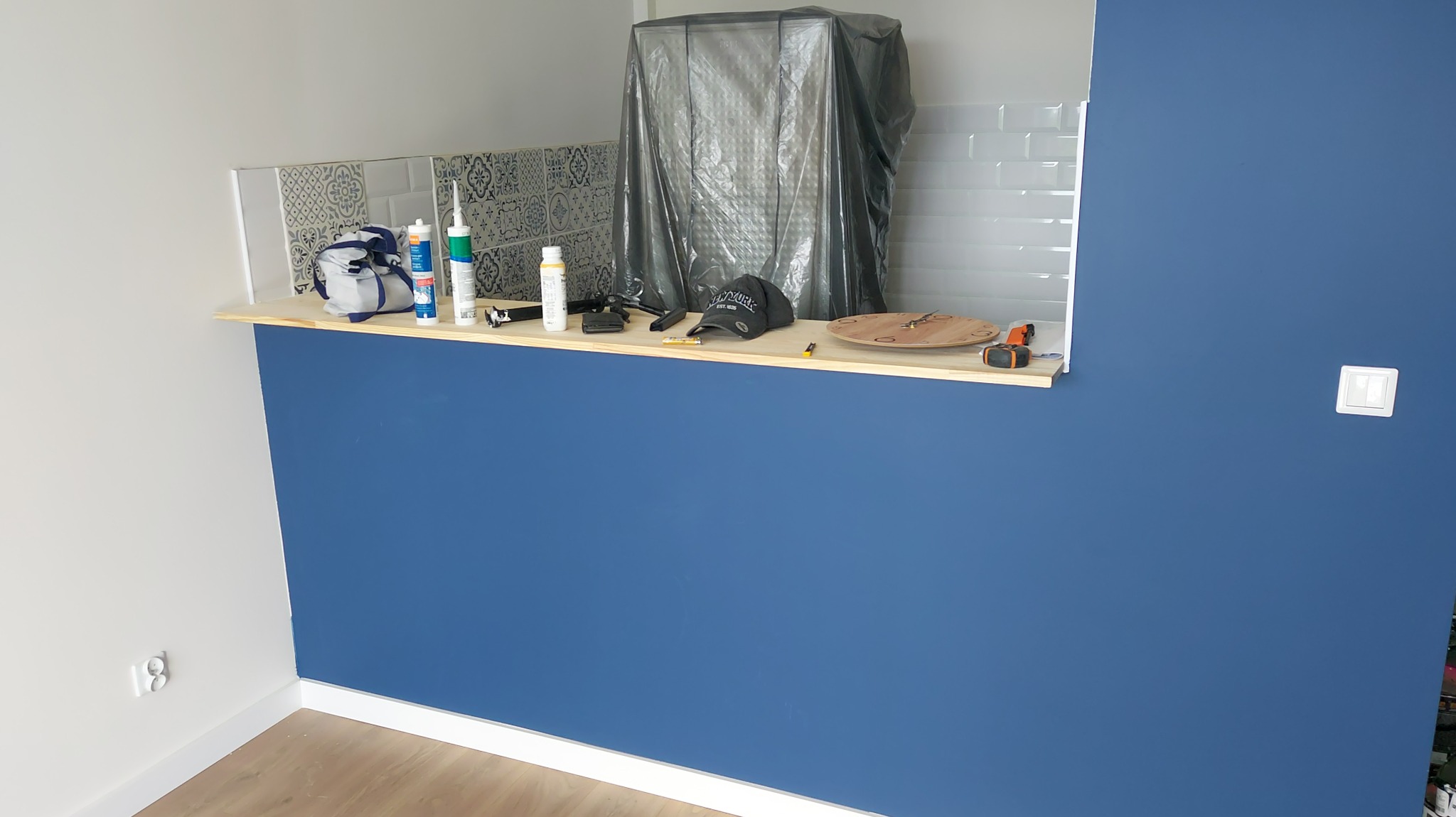 Niebieska ściana po malowaniu z widocznymi narzędziami i materiałami na blacie; fragment kuchni z płytkami dekoracyjnymi.