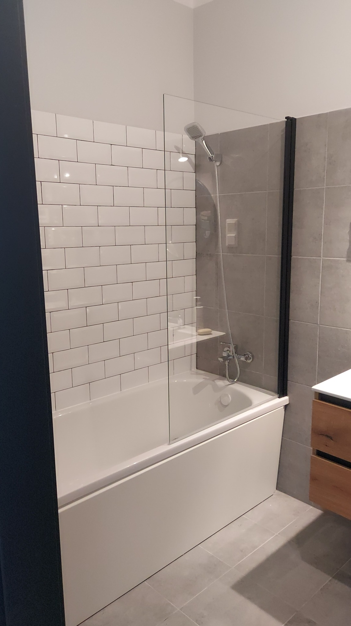 Nowoczesna łazienka z wanną, przeszklonym parawanem z czarną ramą i białą armaturą. Ściany wyłożone białymi i szarymi płytkami. Minimalistyczny design.