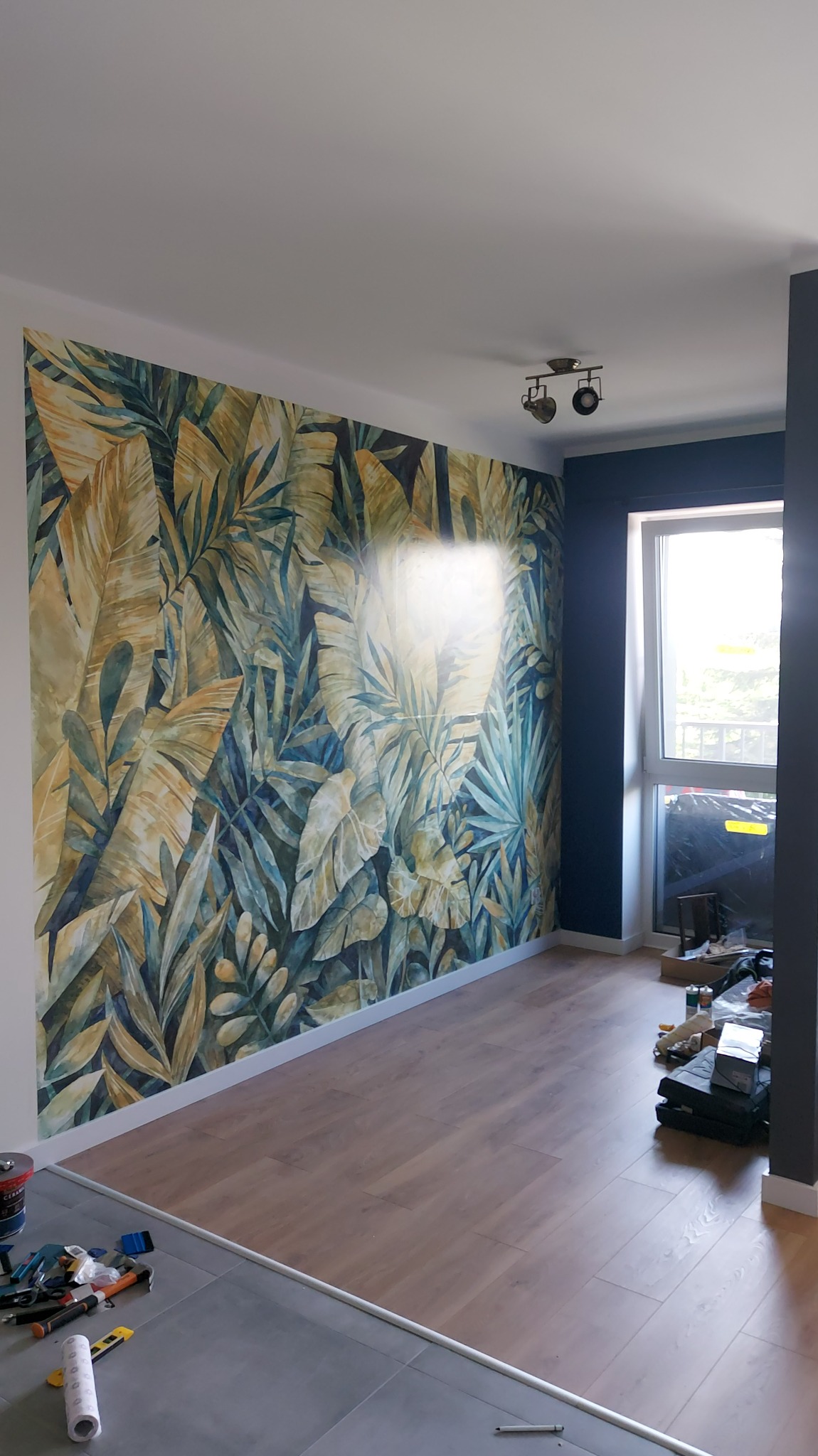 Pokój po remoncie z fototapetą w motywy roślinne, drewnianą podłogą i ciemną ścianą przy oknie. Widoczne narzędzia malarskie.